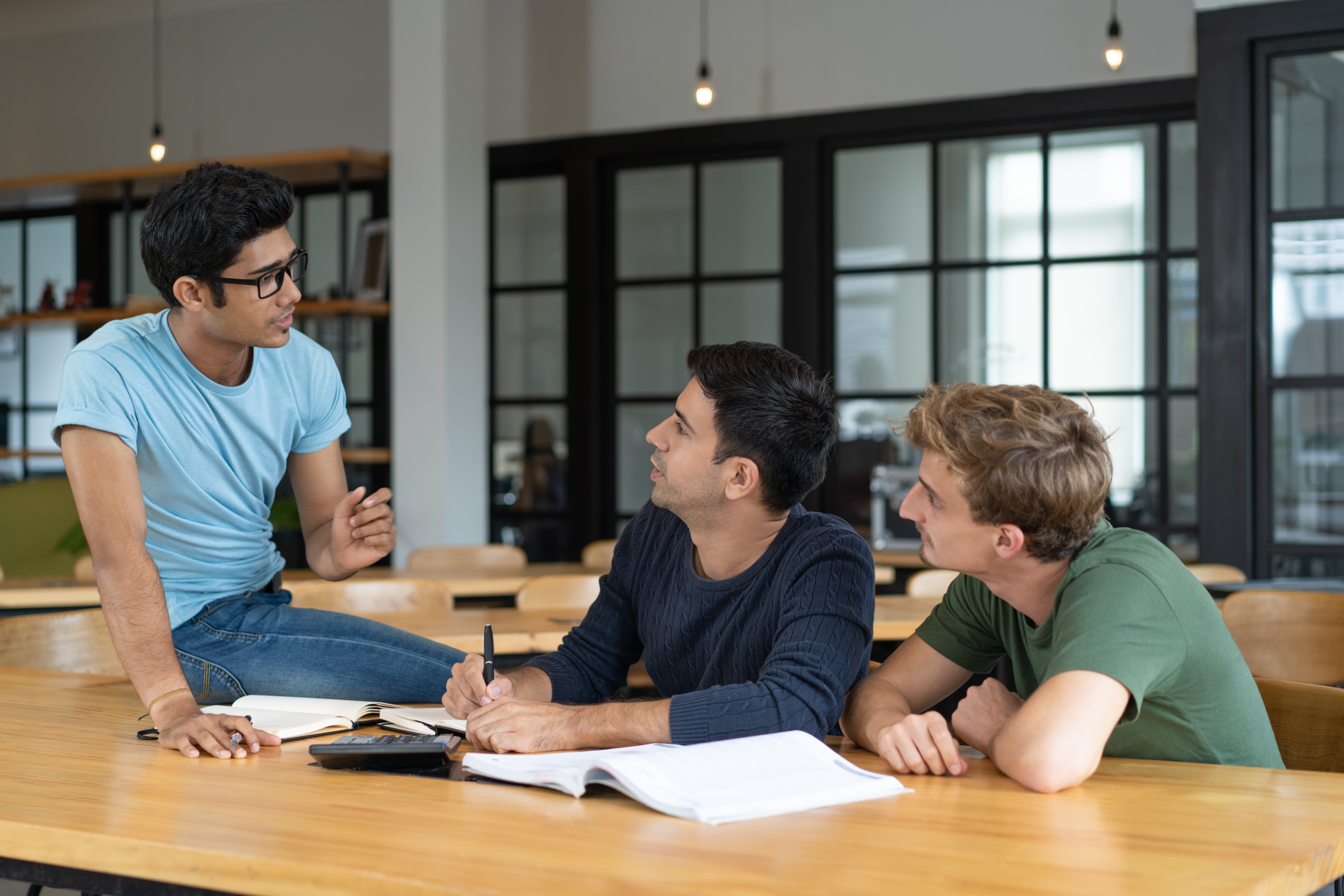 Excited young Indian expert telling team about new product. Three young men arguing at desk with notes and calculator. Corporate communication concept